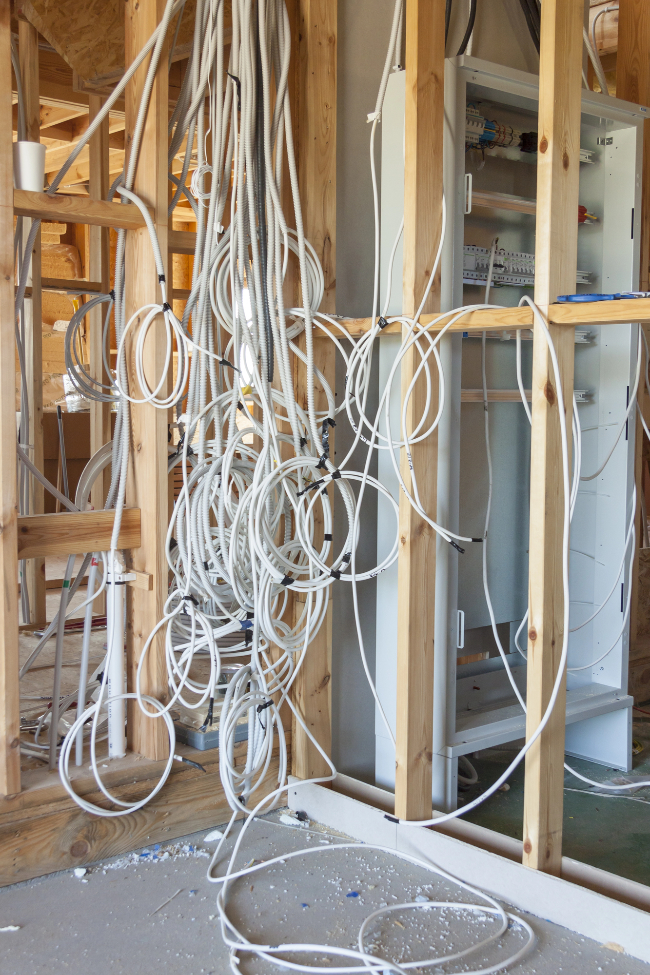 A large number of white electrical cables hang loosely inside a wooden frame of a building under construction, with an open electrical panel visible.