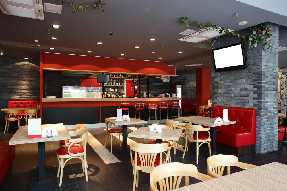 Modern cafe interior with wooden tables, red cushioned seating, and a bar area. Walls are dark gray with brick accents, and there's a TV screen mounted on the wall.